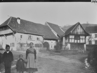 Dorfplatz mit mehreren Gebäuden und Brunnen, links Knabe, kleines Mädchen und Mädchen in Kamera schauend, dahinter dreistöckiges Bauernhaus mit geziegeltem Schleppdach mit Halbwalm, giebelseitige, von Bügen getragene Obergeschosslaube unter Gehrschild, kleiner, traufseitiger Garten mit Lattenzaun, bogenförmiges Tenntor, dahinter Anbau mit erhöhtem, geknicktem Satteldach rechts hinter Brunnen dreistöckiger Stock in Fachwerkbaweise mit  geknicktem Puldach und auskragendem Giebelfeld mit Holzschalung, giebelseite mit Holztor