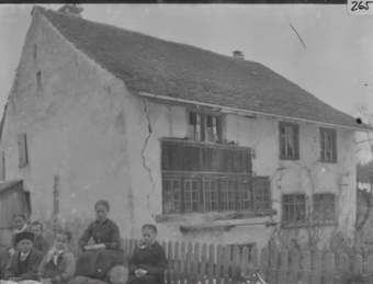 Dreistöckiges, gemauertes Wohnhaus mit geziegeltem Satteldach traufseitige Fensterfront mit Holzrahmen, rechts daneben vertikaler Riss in der Gebäudefassade, im Vordergrund zwei Frauen, zwei Mädchen, ein Knabe und ein Mann vor Lattenzaun sitzend