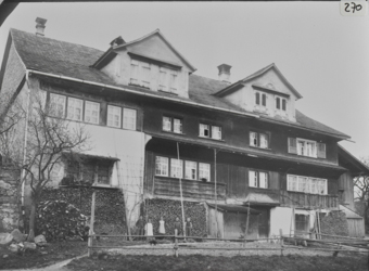 Traufseite vierstöckiges, gemauertes Wohngebäude mit teilweise geschindelter Fassade und Klebdächern, geziegeltes Pultdach mit zwei Giebelgauben, Giebelfeld geschindelt, an Fassade aufgestapeltes Brennholz, davor fünf Kinder stehend, Viehzaun