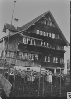 Gieblseite vierstöckiges Wohngebäude mit steilem Schleppdach und Giebelgaube, drei Klebedächer, geschindelte Fassade, Gruppe von Kindern und Frauen hinter Spaliergerüst