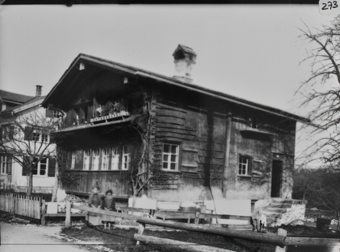 Zweistöckiges, gezimmertes Wohngebäude mit gemauertem Fundament, steinerne Treppe zu erhöhntem, traufseitigem Eingang mit Windfang, giebelseitiges Klebedach, geschindelte Fassade, Kletterpflanze an Hausecke, zwei verschwommene Frauen hinter Bretteraun, im Hintergrund dreistöckiges, gemauertes Wohngebäude