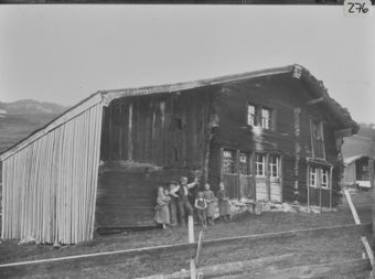 Zweistöckiges Holzhaus mit Schleppdach, links giebelseitiger Anbaumit Holzschalung, vor dem Gebäude posierende Bauernfamilie mit zwei Mädchen, einem Knaben, einem Kleinkind, Mann und Frau, im Vordergrund Bretterzaun