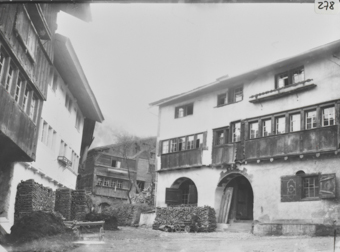 Dorfplatz mit dreistöckigen Gebäuden, rechts gemauertes Wohnhaus mit zwei Kreisbögen im Erdgeschoss, davor Brennholzstapel, im 1. OG Auskragung mit Sprossenfenster und Holzschalung, links hölzerne Auskragung, dahinter gemauertes Wohnhaus mit Brennholzstapel, mitte giebelseite gezimmertes Bauernhaus
