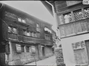 Strasse in Dorf, links Traufseote dreistöckiges Wohnhaus mit gemauertem Fundament und gezimmertem Oberbau, Klebedach über Erdgeschoss, Sprossenfenster und Inschriften im 2. OG, auf dem Fenster hängende Bettwäsche, rechts Fassadenausschnitt mit gemauertem Erdgeschoss und hölzernem Obergeschoss, Butzenscheiben, Brennholzstapel an Hausmauer