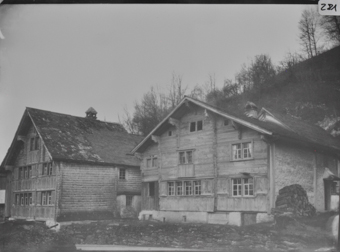 Zwei dreistöckige Holzgebäude mit geschindelten, von Blockkonsolen getragenen Pultdächern und geschindelten traufseitigen Fassaden, gemauerten Fundamenten, im Hintergrund bewaldeter Abhang