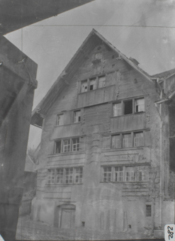 Traufseite sechsstöckiges Gebäude mit gemauertem Fundament und Satteldach 1.&2. OG mit geschindelter Fassade, 3.-6. OG mit Holzschalung, Sprossenfenster, Fensterrähmen mit Holztornamentik, rechts Kletterpflanze, links Ausschnitt Holztor