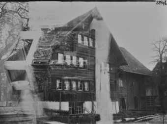 Doppelaufnahme, vierstöckiges Wohnhaus mit geziegeltem Satteldach Erdgeschoss gemauert, Oberbau gezimmert, Giebelseite mit Blockkonsolen, Sprossenfenstern und Friesen, davor Ziergarten mit Spaliergestell und Lattenzaun, rechts im Hintergrund Ökonomieteil mit Holzschalung und geziegeltem Dach, zweite Aufnahme von Kachelofen überlagert