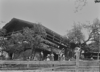 Mehrstöckiges Bauernhaus aus Mischbau mit Fundament aus Stein, Satteldach und Balkon, diverse Sträucher und Bäume verdecken den vorderen Teil des Hauses, im Garten sthen diverse Personen, Gartenzaun grenzt Garten ein, neben dem Bauernhaus rechts ist ein weiteres Haus ersichtlich