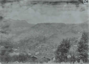 Ansicht eines Dorfes mit mehreren Häusern, vorne sind belaubte Bäume auf einer Wiese, im Hintergrund Hügelkette