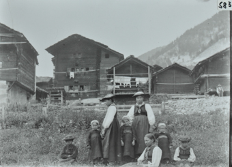 Drei Frauen und Kinder posieren auf der Wiese mit hohem Gras, am Ende der Wiese ein Holzzaun sowie eine Steinmauer, dahinter sind mehrere mehrstöckige Wohnhäuser mit Fundament aus Stein, Satteldächer auf den Häusern