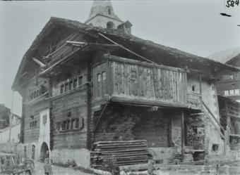 Dreistöckiges Gebäude mit gemauertem Fundament, Satteldach, Steintreppe führt zu einem Balkonähnlichen Eingang, unter dem Balkon gestapeltes Holz, hinter dem Haus ragt ein Kirchenturm hervor, vor dem Haus führt Strasse vorbei
