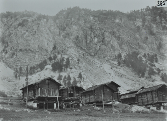 Ansicht des Dorfes mit einer Wiese und mehreren Gebäuden aus Holz, Gelände abfallend, die Gebäude stehen auf einem Fundament aus Stein, Satteldächer, Bergkette im Hintergrund