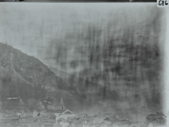 Ansicht des Dorfes aus der Vogelperspektive, mehrere Gebäude und ein Kirchenturm ersichtlich, Hügel im Hintergrund