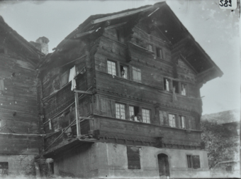 Vierstöckiges Wohnhaus mit Fundament aus Stein, Satteldach, zwei Personen schauen aus dem Fenster, links neben dem Fenster befindet sich weiterer Anbau