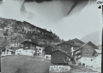 Ansicht des Dorfes in einer Hanglage, mehrere mehrstöckige Häuser aus Holz mit Satteldach, teilweise auf Fundament aus Stein, Dorfstrasse führt durch Dor, im Hintergrund Hügel
