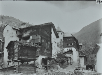 Mehrstöckiges Wohnhaus mit Satteldach, Fundament aus Steinen, ein Teil der Fassade aus Holz ein Teil gemauert, Satteldach, hinterer Bereich weiterer Anbau aus Steinen, Strasse führt neben das Gebäude durch das Dorf, im Vordergrund ein Speicher aus Holz mit Satteldach