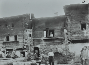 Menschengruppe posiert, teils sitzend, vor einem Mehrfach-Wohnhaus, weitere pesonen schauen aus den Fenster, Gebäude mit Fundament aus Stein, oberer Teil aus Holz, jeweils mit Satteldach