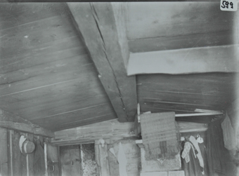 Innenaufnahme eines Zimmers mit einer Decke aus Holz, Holzbalken verläuft längst durch die Decke, hinteren Ende sind diverse Kleider sowie ein Tuch aufgehängt