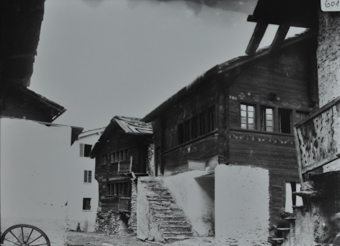 Ansicht eines Teils des Dorfs, Strasse führt duch das Dorf, dahinter befinden sich zwei Wohnhäuser aus Holz, das Fundament ist jeweils aus Stein, Steintreppe führt zu den Eingängen, Satteldach