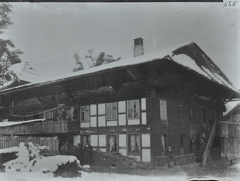 Zweistöckiges Bauernhaus aus Holz, Walmdach, mehrere Personengruppen posieren vor dem Haus, auf der Holztreppe und auf dem Balkon, vor dem Haus liegt gestapeltes Brennholz, Umgebung verschneit