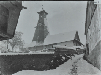 Ansicht der Frühmesskapelle aus Steinbau, hoher Kirchenturm mit Uhr und Glocken, eine Leiter führt vom Boden zur Turmuhr, ein Weg mit einerm Holzzaun führt zur Kirche, Umgebung verschneit