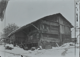 Zweistöckiges Bauernhaus aus Holz mit Schleppdach, wenige Fenster vorhanden, diverse Gegenstände liegen vor und neben dem Haus, Umgebung verschneit