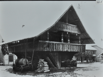 Zweistöckiges Bauernhaus aus Holz mit Satteldach, zwei Balkons, unter dem Haus liegt gestapeltes Brennholz sowie ein Holzkarren mit einem Fass, weitere Gebäude im Hintergrund, Umgebung verschneit