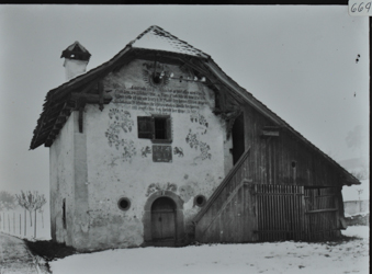 Zweistöckiges Gebäude, teils gemauert und teils Holzbau, Holztreppe führt zur oberen Etage, Krüppelwalmdach, Inschrift und Zeichnungen auf der Fassade, Strasse führt am Haus vorbei, Umgebung verschneit