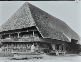 Mehrstöckiges Bauernhaus aus Holz mit Krüppelwalmdach, Garten vor dem Haus, Brunnen sowie Holzkarren neben dem Haus, Strasse führt neben Haus vorbei