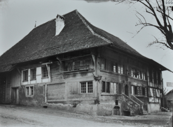 Zweistöckiges Gebäude aus Holz und gemauerten Fundament, Steintreppe führt zum Eingang, Gelände leicht abfallend, Strasse führt am Haus vorbei