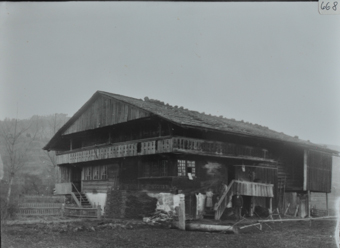 Zweistöckiges Wohnhaus aus Holz mit gemauerten Fundament, Satteldach und Balkon, zwei Treppen führen zu den Eingängen, Menschen posieren vor und auf der Treppe, gestapeltes Brennholz vor dem Haus
