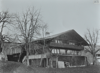 Zweistöckiges Bauernhaus aus Holz, Satteldach und Balkon, vor und neben dem Haus liegt gestapeltes Brennholz, vor dem Haus sind kahle Bäume, am hinteren Teil ist Scheune angebaut