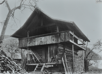 Zweistöckiges Gebäude aus Holz, Schleppdach, Balkon unter dem Dach, kleine Treppe führt Eingang, neben, vor und hinter dem Haus gestapeltes Brennholz, Holzschlitten an Haus angelehnt, kahle Bäume umd das Haus 