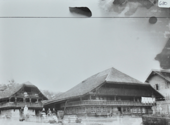 Dreistöckiges Bauernhaus aus Holz, Krüppelwalmdach, auf oberen Etagen Balkon, weitere Wohnhäuser in der Umgebung, vor den Gebäuden posieren Frauen, eine Strasse führt vor den Häusern vorbei