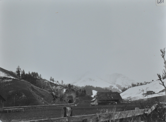 Mehrstöckiges Bauernhaus aus Holz auf einer Wiese, Krüppelwalmdach, Zugschienen führen am Haus vorbei in ein Tunnel, im Hintergrund verschneite Berge
