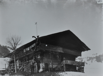 Zweistöckiges Bauernhaus aus Holz mit Fundament aus Holz, Satteldach, Holztreppe führt zur ersten Etage wo eine Frau posiert, weitere Person posiert auf der Wiese vor dem Haus, gestapeltes Brennholz neben dem Haus, kahler Baum neben dem Haus, Umgebung verschneit