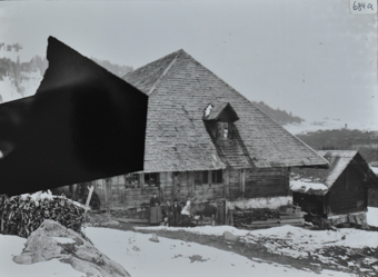 Bauernhaus aus Holz und Walmdach, eine Frau, Kinder sowie eine Kuh posieren vor dem Haus, Brennholz liegt unter und neben dem Haus, rechts neben dem Haus befindet sich ein weiters Gebäude, Umgebung verschneit