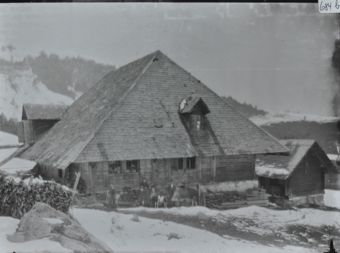 Bauernhaus aus Holz und Walmdach, eine Frau, Kinder sowie eine Kuh posieren vor dem Haus, Brennholz liegt unter und neben dem Haus, rechts neben dem Haus befindet sich ein weiters Gebäude, Umgebung verschneit