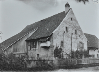 Zweistöckiges gemauertes Wohnhaus mit Satteldach und einem erweiterten Schleppdach, Kletterpflanzen an der vorderzeite der Fassade, Gartenzaun aus Holz um das Haus, weiteres Gebäude hinter dem Wohnhaus, Strasse führt vor dem Haus vorbei