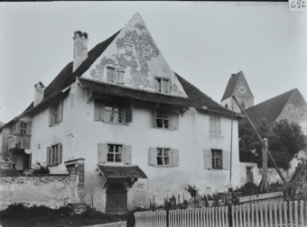 Mehrstöckiges gemauertes Wohnhaus, Fusswalmdach, kleines Dach oberhalb der Eingangstür, Steinmauer links und rechts neben dem Haus, Strasse vor dem Haus, hinten rechts weiteres Gebäude