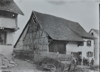 Mehrstöckiges Bauernhaus, grösstenteils gemauert, eine Wand aus Holz, Schleppdach, Menschengruppe posiert vor dem Hauseingang, im Vordergrund ein kleiner Weg sowie ein Holzaun mit Heu und Schubkarre