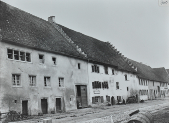 Aneinander gereihte Wohnhäuser, drei- und zweistöckig, alle mit Satteldach, Strasse führt beim Haus vorbei, zwei Kinder posieren vor dem Haus, Holzfass im Vordergrund