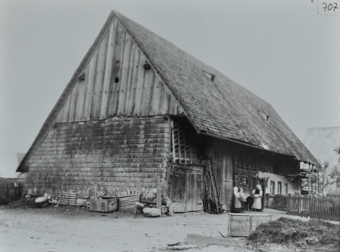Mehrstöckiges Bauernhaus, Haus teils gemauert und teils aus Holz, Satteldach, vorderer Teil des Gebäudes als Scheune genutzt, zwei Frauen stehen vor dem Haus, vor dem Haus Garten mit Gartenzaun, Misthaufen im Vordergrund