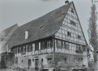 Mehrstöckiges Bauernhaus, Parterre aus Stein, oberer Bereich gemauert, steiles Satteldach, Holzkarren vor dem Haus, hinter dem Haus befindet sich ein weitere Anbau