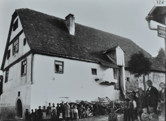 Mehrstöckiges Gebäude gemauert, Zwerchdach, Strasse verläuft neben dem Gebäude, vor und hinter der Strasse posieren Erwachsene und Kinder, vor dem Haus liegt gestapeltes Brennholz