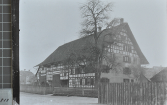 Mehrstöckiges gemauertes Bauernhaus mit Satteldach, Fassade mit Holzelementen verziert, grosser kahler Baum steht im Garten vor dem Haus, Holzzaun rund um den Garten, ein Weg führt seitlich des Hauses vorbei