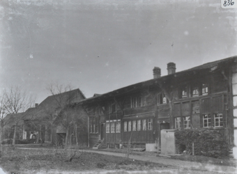 Zweistöckiges Mehrfach-Wohnhaus mit Fundament aus Stein, Etagen aus Holz, Satteldach, welches mit Holzbalken an den Enden befestig ist, Holztreppe führt zum Eingang, gestapeltes Brennholz vor dem Haus, Weg führt vor dem Gebäude vorbei, links neben den Gebäude weitere Gebäude, kahle Bäume und Sträucher vor dem Gebäude
