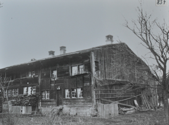 Zweistöckiges Baurnhaus mit gemauerten Fundament, Etagen aus Holz, Satteldach mit Kaminen, Holztreppe vor dem Eingang, gespapeltes Brennholz vor dem Haus, an der rechten Hauswand liegen Holztreppen und weitere Holzteile, kahler Baum im Vorgarten