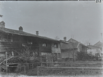 Zweistöckiges Bauernhaus aus Holz mit Satteldach, mehrere Haufen gestapeltes Brennholz sowie Rasen vor dem Haus, weitere Gebäude im Hintergrund