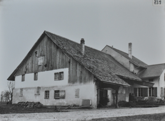 Mehrstöckiges Bauernhaus, unteren zwei Etagen gemauert, oberer Teil aus Holz, Schleppdach, weiterer gemauerterer Anbau am hinteren Ende, vor dem Gebäude liegt ein Steinhaufen, Weg führt neben dem Gebäude vorbei